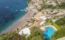 Aerial view of Positano, Italy, showcasing the beach, colorful hillside buildings, and a swimming pool in the foreground, with boats in the turquoise sea.