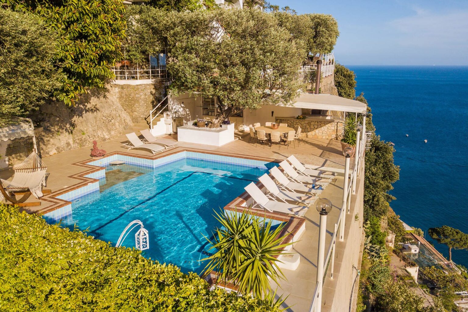 Luxurious cliffside villa with a stunning infinity pool overlooking the ocean, surrounded by lush greenery and outdoor seating. Perfect for a relaxing getaway. | Only on SopranoVillas