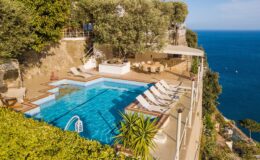 Luxurious cliffside villa with a stunning infinity pool overlooking the ocean, surrounded by lush greenery and outdoor seating. Perfect for a relaxing getaway.