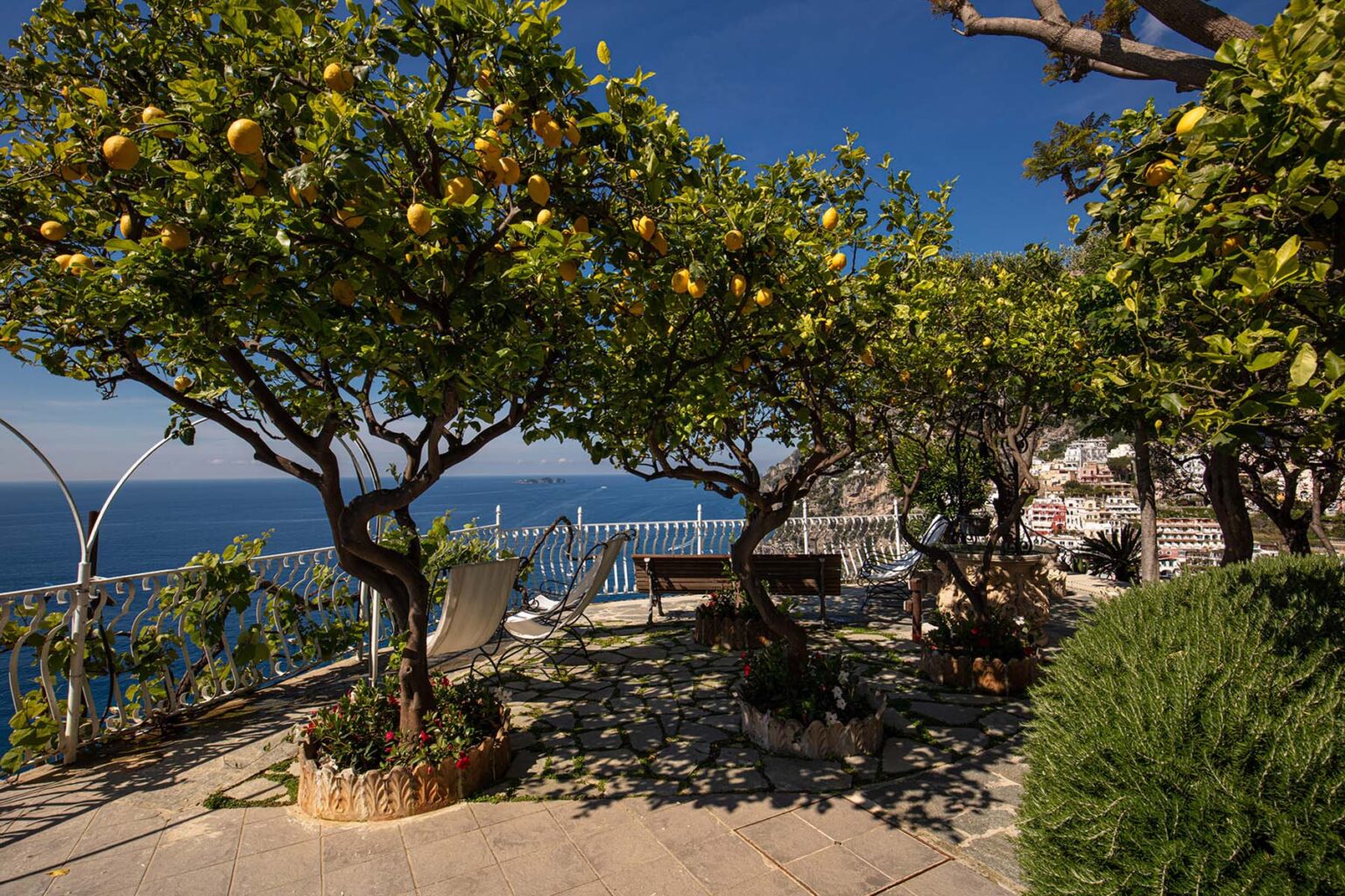 Lush lemon trees in a coastal garden overlooking the sea, featuring a relaxing seating area and bright blue sky, perfect for outdoor relaxation and scenic views. | Only on SopranoVillas
