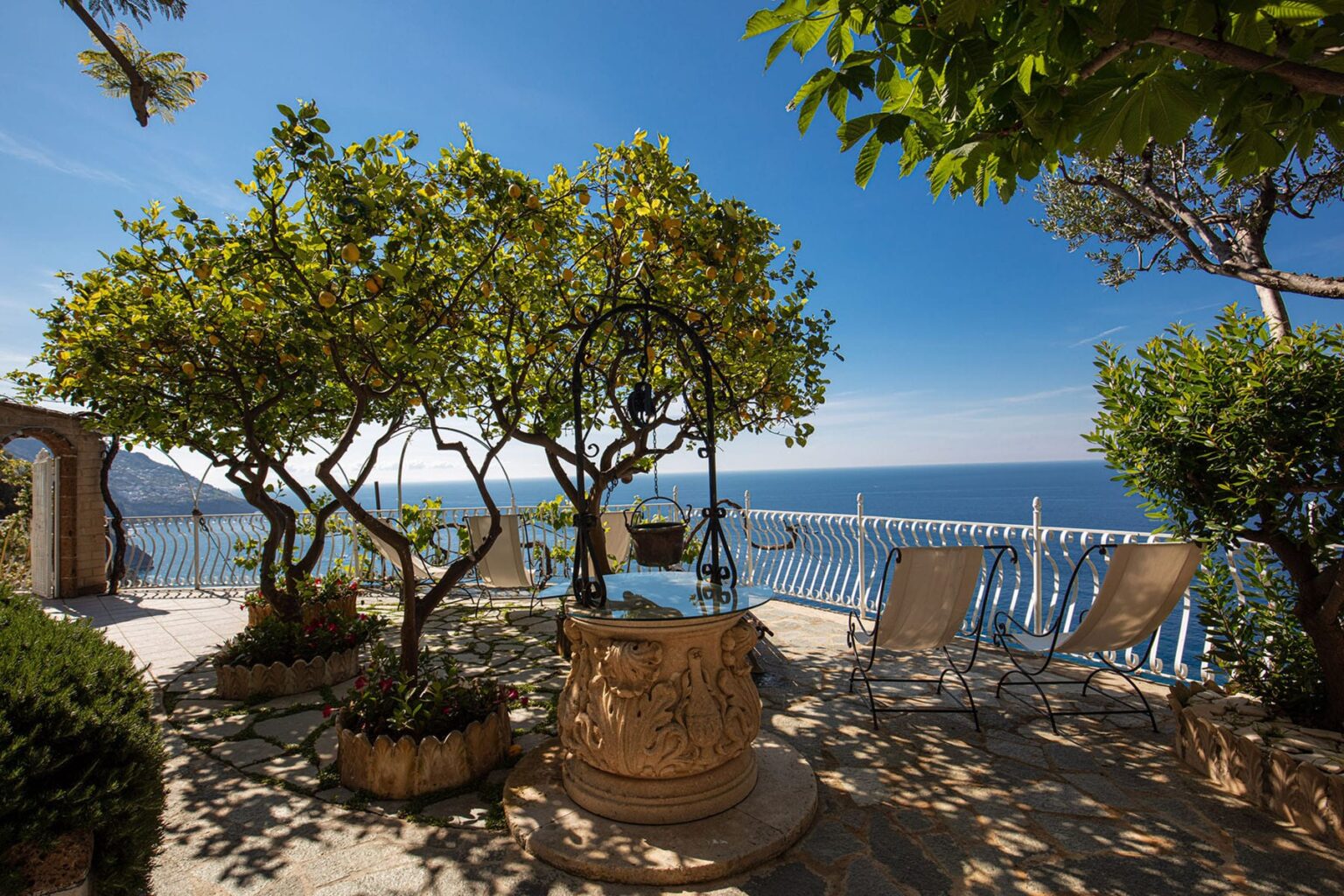 Scenic outdoor terrace with a stone well, lush greenery, and lounge chairs overlooking a beautiful sea view under a clear blue sky. | Only on SopranoVillas