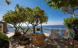 Scenic outdoor terrace with a stone well, lush greenery, and lounge chairs overlooking a beautiful sea view under a clear blue sky.