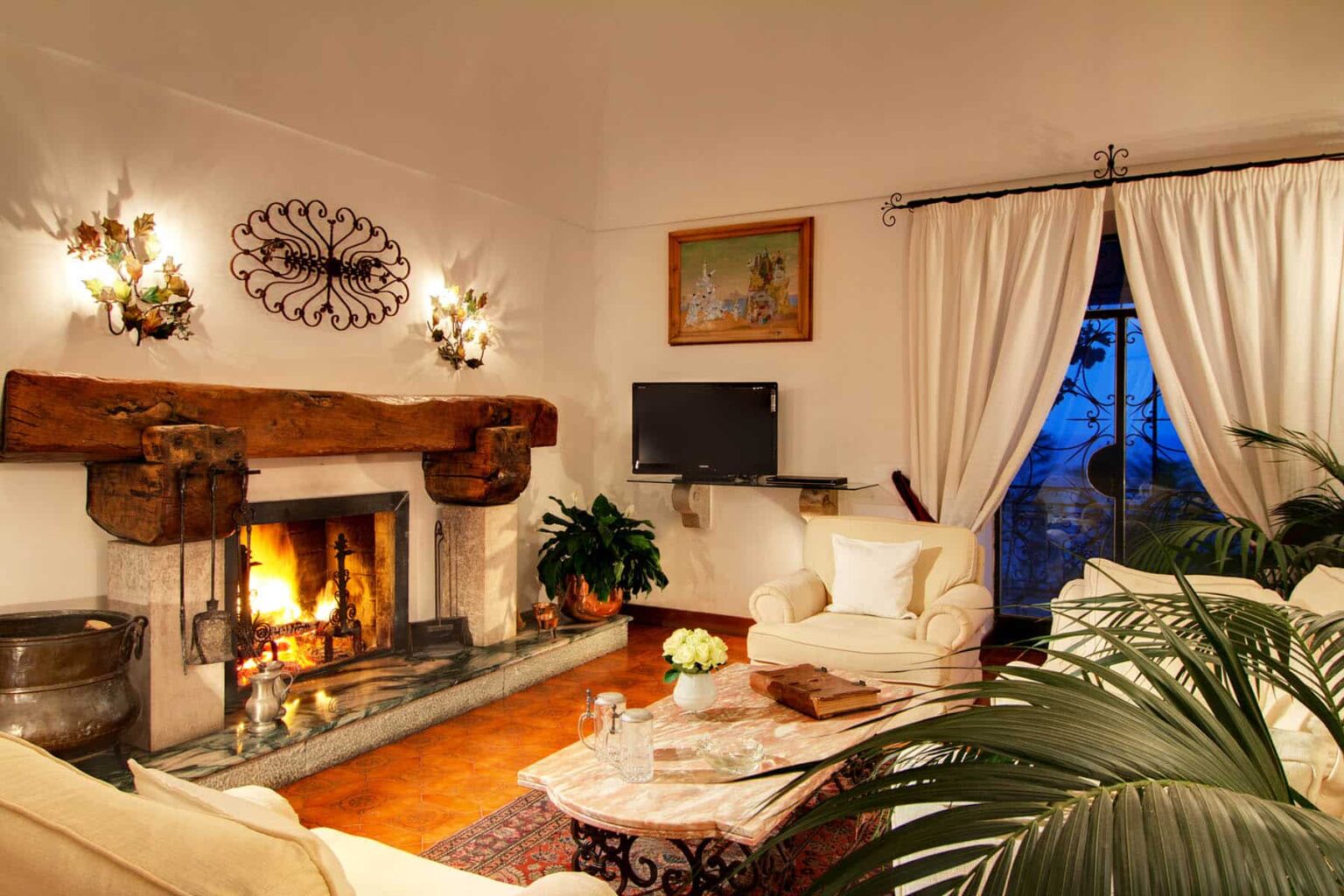 Cozy living room with a fireplace, white sofas, and elegant decor, featuring large windows with sheer curtains and a flat-screen TV, creating a warm and inviting atmosphere. | Only on SopranoVillas