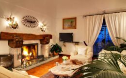 Cozy living room with a fireplace, white sofas, and elegant decor, featuring large windows with sheer curtains and a flat-screen TV, creating a warm and inviting atmosphere.