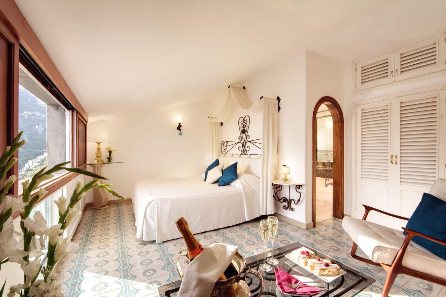 Bright and airy hotel room featuring a queen-sized bed, elegant decor, and large windows offering scenic views. The space includes a cozy seating area, stylish furnishings, and natural light enhancing the tranquil atmosphere. Ideal for relaxation and comfort during a getaway. | Only on SopranoVillas