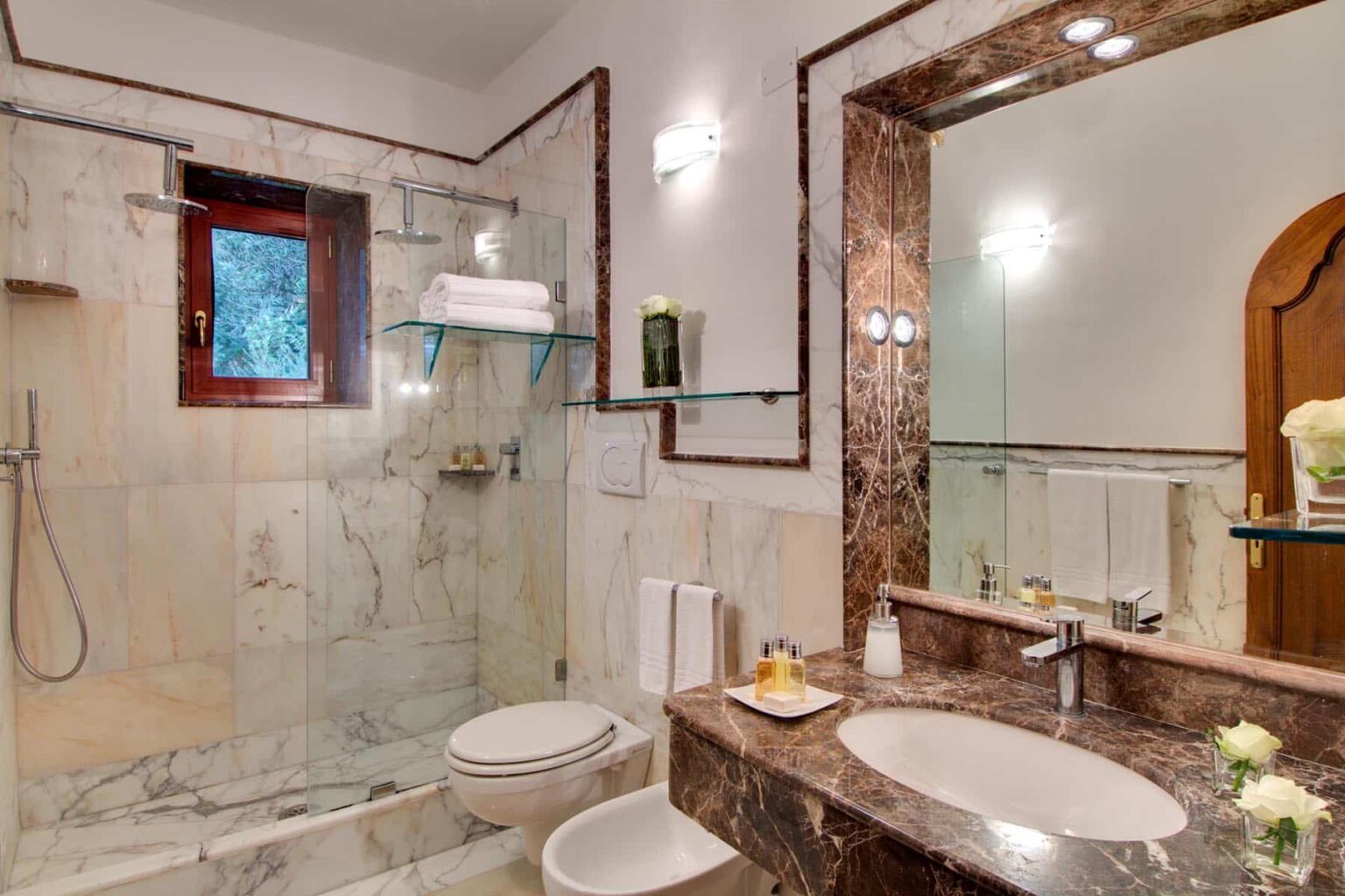 Modern bathroom featuring marble walls, a glass shower, a large mirror, and elegant fixtures, with natural light entering through a small window. | Only on SopranoVillas
