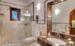 Modern bathroom featuring marble walls, a glass shower, a large mirror, and elegant fixtures, with natural light entering through a small window.