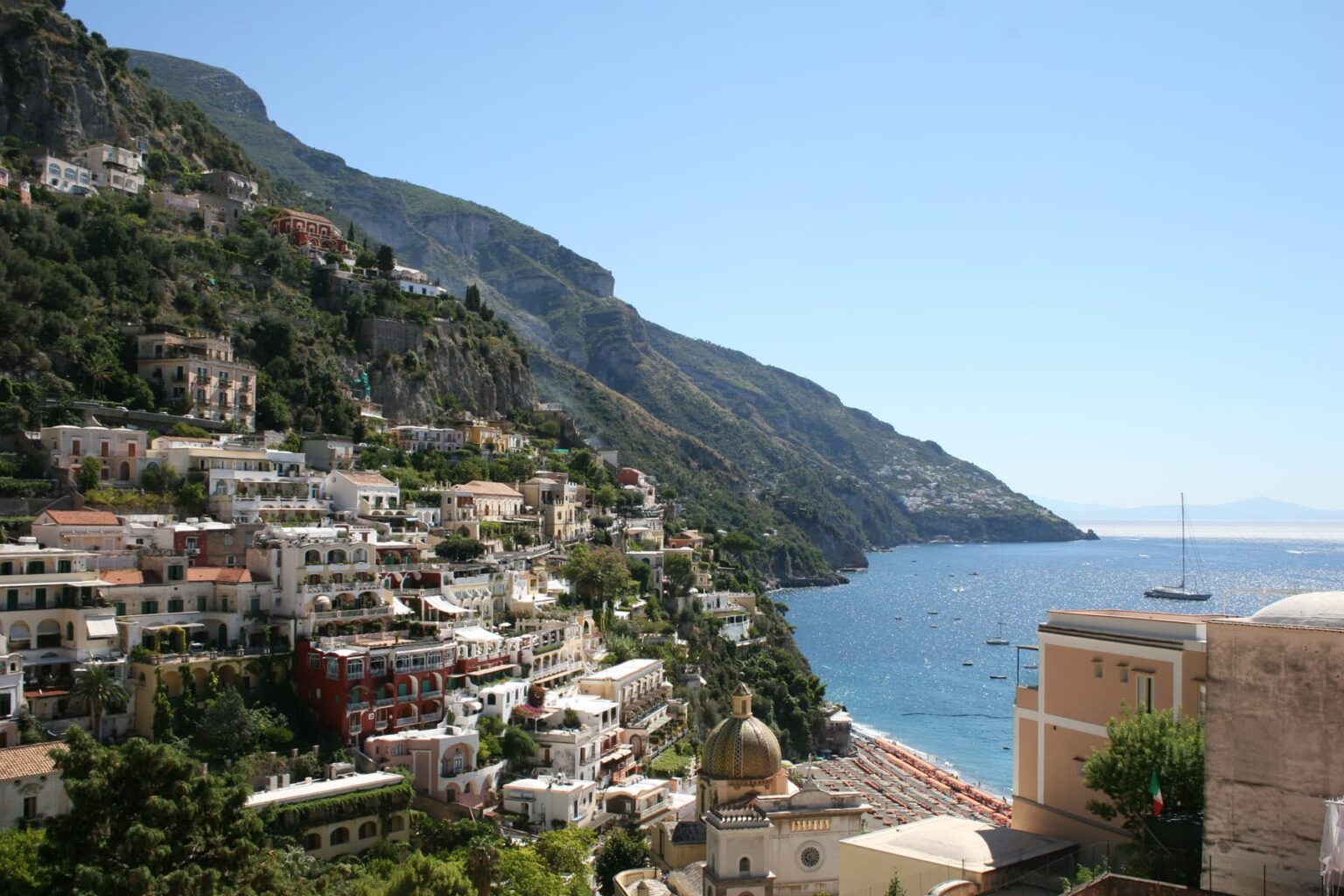 Panoramic view of Positano, Italy, showcasing colorful cliffside houses against a backdrop of mountains and the Mediterranean Sea under a clear blue sky. Ideal for travel and tourism content. | Only on SopranoVillas