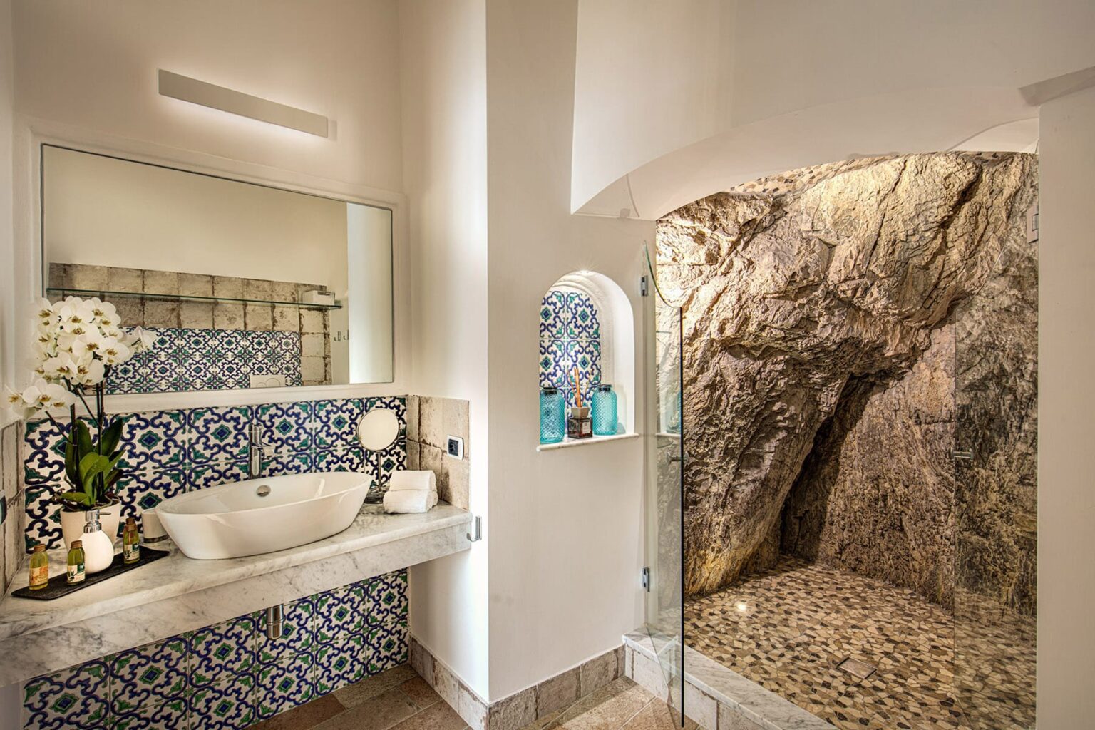 Luxurious bathroom featuring a modern sink, intricate blue patterned tiles, and a unique rock wall shower area, complemented by elegant decor and natural lighting. | Only on SopranoVillas