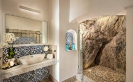 Luxurious bathroom featuring a modern sink, intricate blue patterned tiles, and a unique rock wall shower area, complemented by elegant decor and natural lighting.