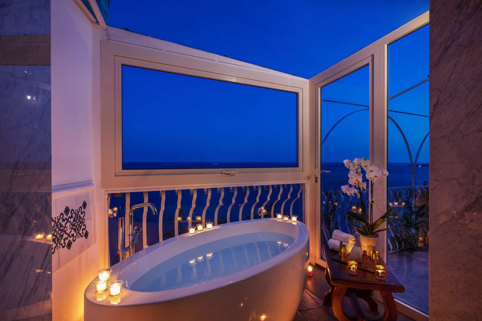 Luxurious bathroom with a freestanding bathtub, surrounded by candles and an ocean view at dusk, featuring large windows that open to a serene outdoor space. | Only on SopranoVillas