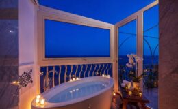 Luxurious bathroom with a freestanding bathtub, surrounded by candles and an ocean view at dusk, featuring large windows that open to a serene outdoor space.