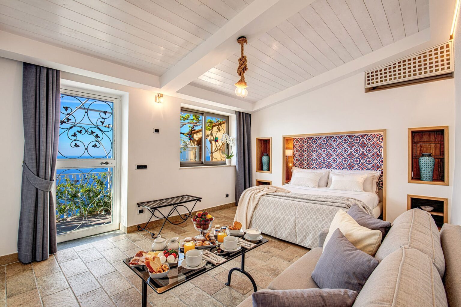 Cozy hotel room featuring a king-sized bed, elegant decor, and a view of the sea through large windows. The space includes a comfortable sofa and a coffee table set with breakfast items, creating a welcoming atmosphere. | Only on SopranoVillas