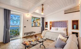 Cozy hotel room featuring a king-sized bed, elegant decor, and a view of the sea through large windows. The space includes a comfortable sofa and a coffee table set with breakfast items, creating a welcoming atmosphere.
