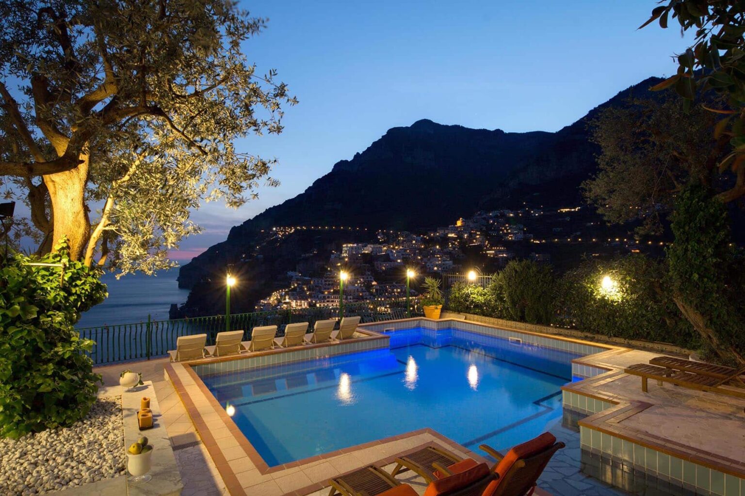 Luxurious villa with a serene swimming pool at dusk, surrounded by lush greenery and illuminated by soft lights, overlooking a picturesque hillside village. | Only on SopranoVillas