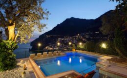Luxurious villa with a serene swimming pool at dusk, surrounded by lush greenery and illuminated by soft lights, overlooking a picturesque hillside village.