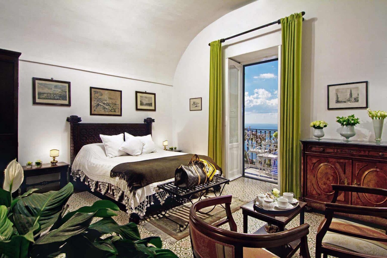 Cozy bedroom with a large bed, green curtains, and a scenic view through an open door, featuring elegant decor, artwork on the walls, and a wooden cabinet with fresh flowers. | Only on SopranoVillas