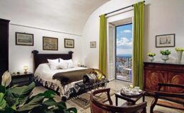 Cozy bedroom with a large bed, green curtains, and a scenic view through an open door, featuring elegant decor, artwork on the walls, and a wooden cabinet with fresh flowers.
