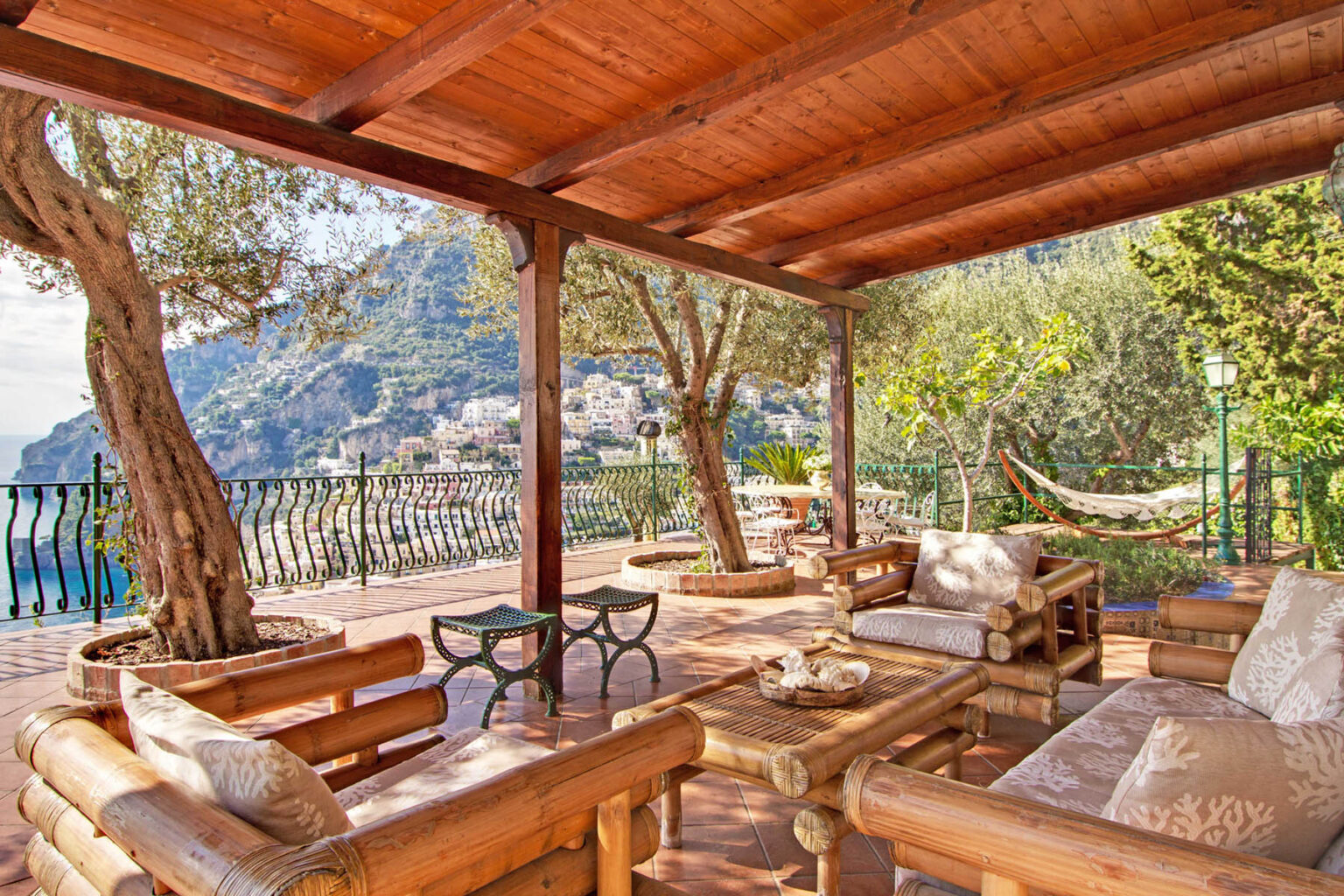 Cozy outdoor seating area with wooden furniture under a pergola, surrounded by lush greenery and a scenic view of hills and coastline. Perfect for relaxation and enjoying nature. | Only on SopranoVillas