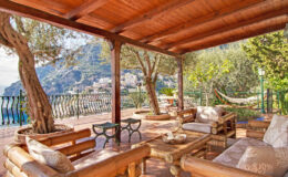 Cozy outdoor seating area with wooden furniture under a pergola, surrounded by lush greenery and a scenic view of hills and coastline. Perfect for relaxation and enjoying nature.