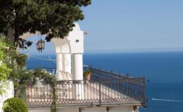 Scenic ocean view from a balcony with white architecture and lush greenery, overlooking a calm blue sea.