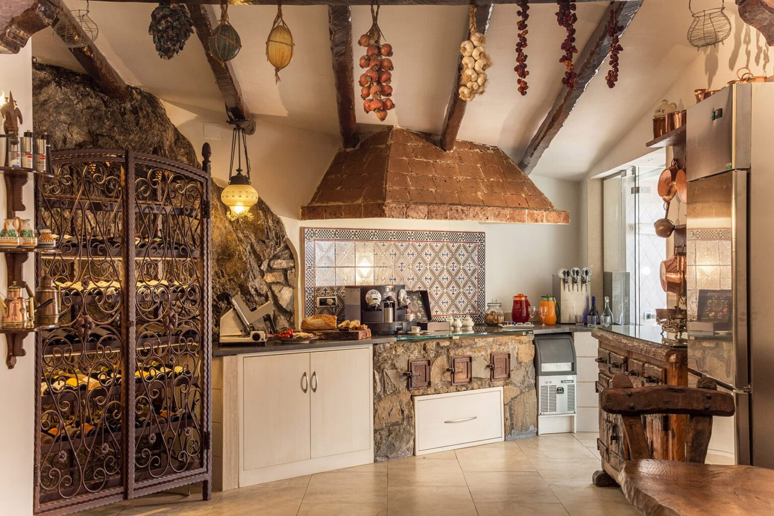 Rustic kitchen interior featuring exposed wooden beams, a stone wall, and a decorative range hood. The space includes a mix of modern appliances and traditional decor, with hanging herbs and spices adding a warm, inviting atmosphere. | Only on SopranoVillas