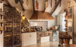 Rustic kitchen interior featuring exposed wooden beams, a stone wall, and a decorative range hood. The space includes a mix of modern appliances and traditional decor, with hanging herbs and spices adding a warm, inviting atmosphere.