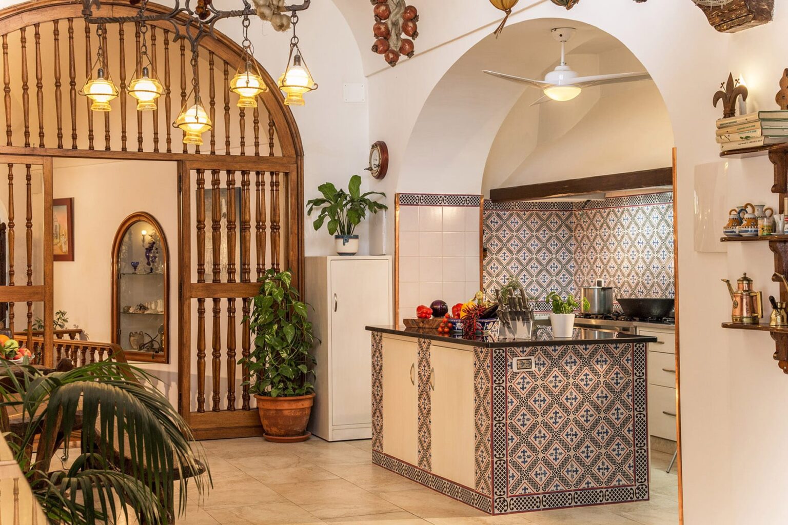Bright and airy kitchen interior featuring a decorative archway, tiled backsplash, and a central island adorned with fresh fruits. Lush greenery and warm lighting enhance the inviting ambiance, showcasing a blend of modern and traditional design elements. | Only on SopranoVillas