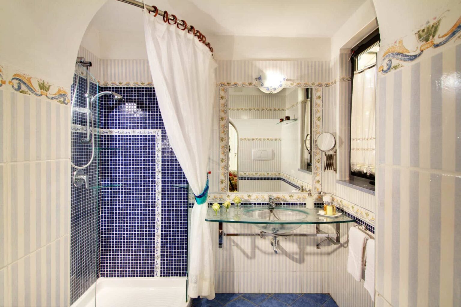 Elegant bathroom featuring a blue tiled shower, a large mirror, and decorative accents. The space includes a glass sink and stylish fixtures, creating a modern yet classic ambiance. | Only on SopranoVillas