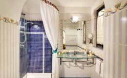 Elegant bathroom featuring a blue tiled shower, a large mirror, and decorative accents. The space includes a glass sink and stylish fixtures, creating a modern yet classic ambiance.