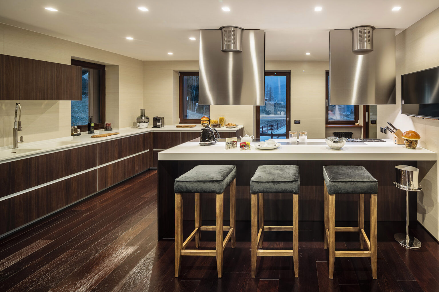 Modern kitchen interior featuring sleek wooden cabinetry, a large island with bar stools, stainless steel appliances, and ample natural light from large windows. The design emphasizes a contemporary aesthetic with dark wood flooring and minimalist decor. | Only on SopranoVillas