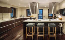 Modern kitchen interior featuring sleek wooden cabinetry, a large island with bar stools, stainless steel appliances, and ample natural light from large windows. The design emphasizes a contemporary aesthetic with dark wood flooring and minimalist decor.