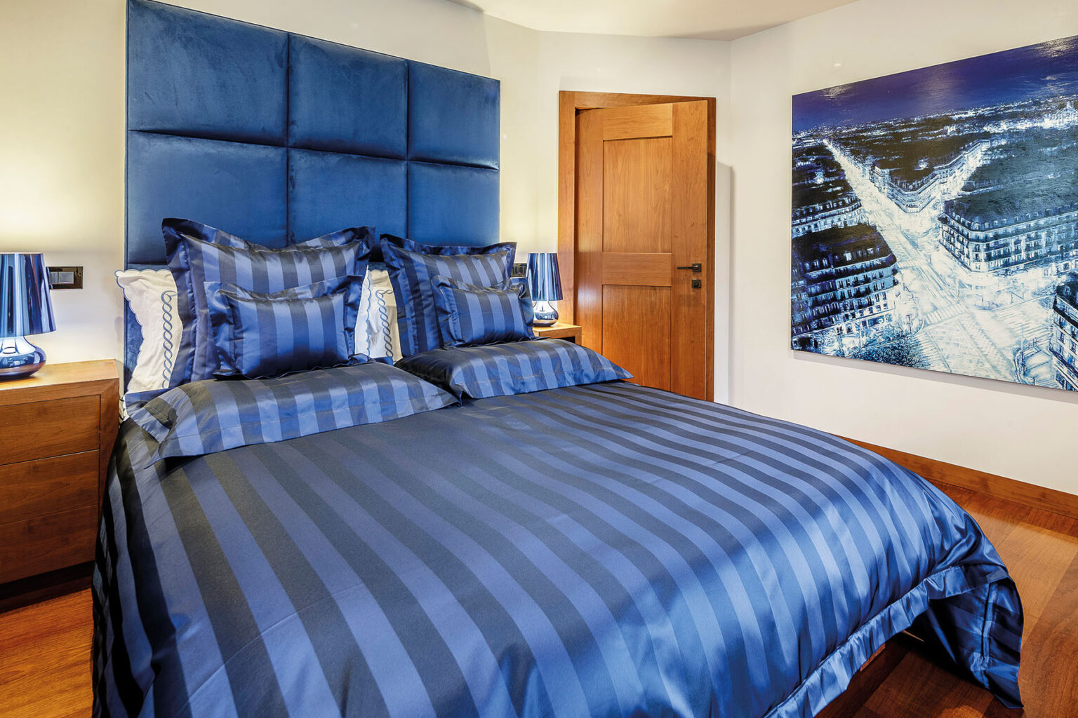 Modern bedroom featuring a luxurious blue striped bedding set, plush pillows, and a velvet headboard. A stylish wooden bedside table with a blue lamp complements the decor. The room includes a wooden door and a large artwork depicting a cityscape at night. | Only on SopranoVillas
