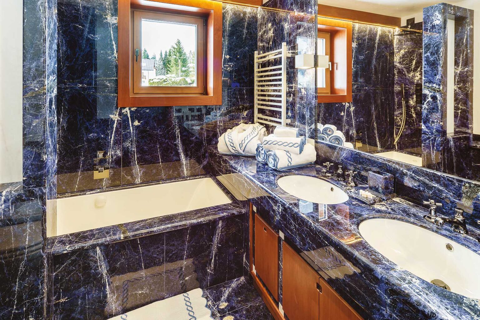 Luxurious bathroom featuring elegant blue marble walls and countertops, a spacious bathtub, dual sinks with modern fixtures, and neatly arranged towels, creating a sophisticated and stylish ambiance. Natural light streams in through a window, enhancing the overall aesthetic. | Only on SopranoVillas