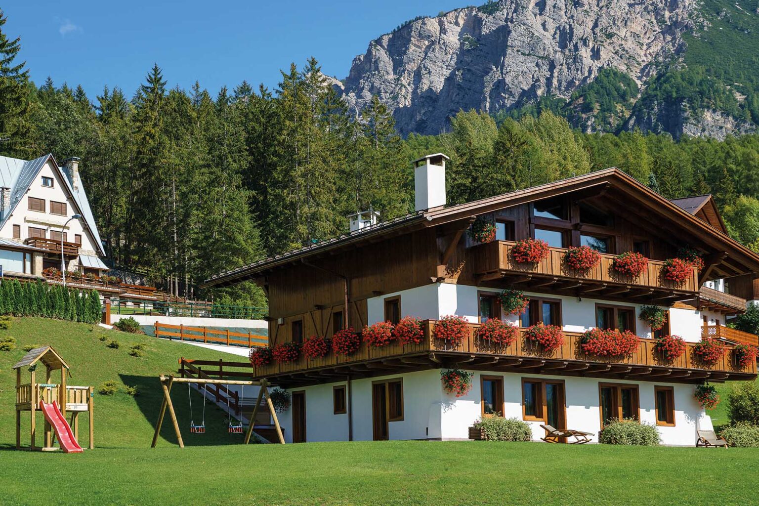 Scenic alpine chalet with flower-filled balconies, surrounded by lush greenery and mountains, featuring a children's play area with a slide and swings. Ideal for family getaways and nature retreats. | Only on SopranoVillas