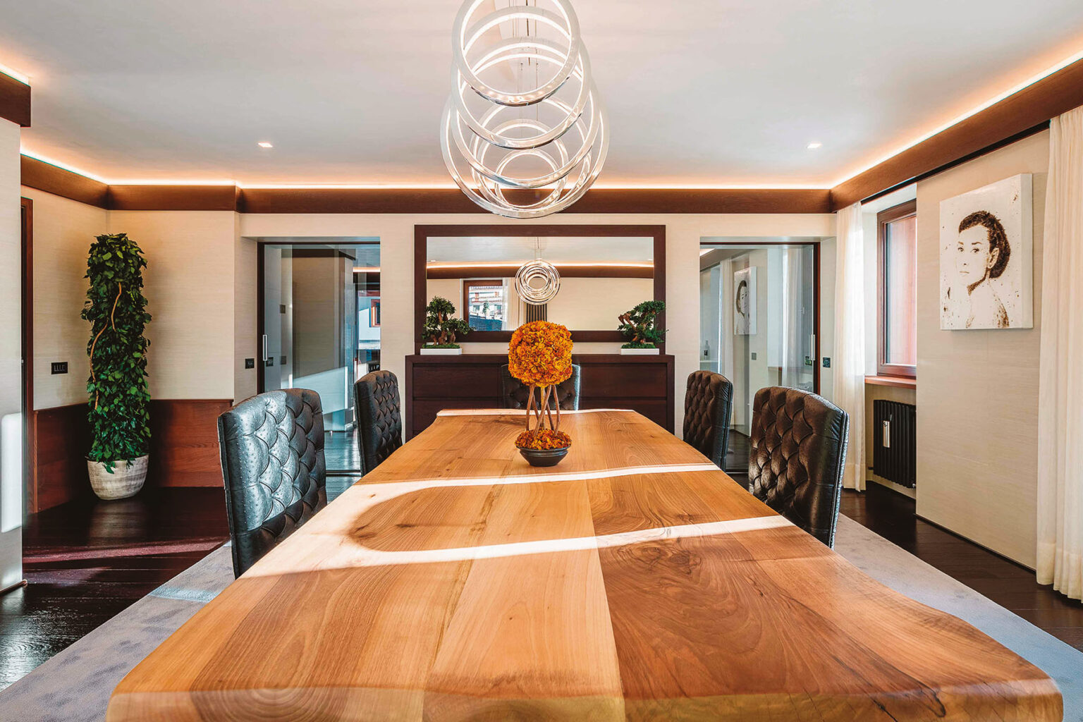 Modern dining room featuring a large wooden table, elegant leather chairs, and contemporary lighting fixtures. The room is accentuated with decorative plants and artwork, creating a stylish and inviting atmosphere. | Only on SopranoVillas