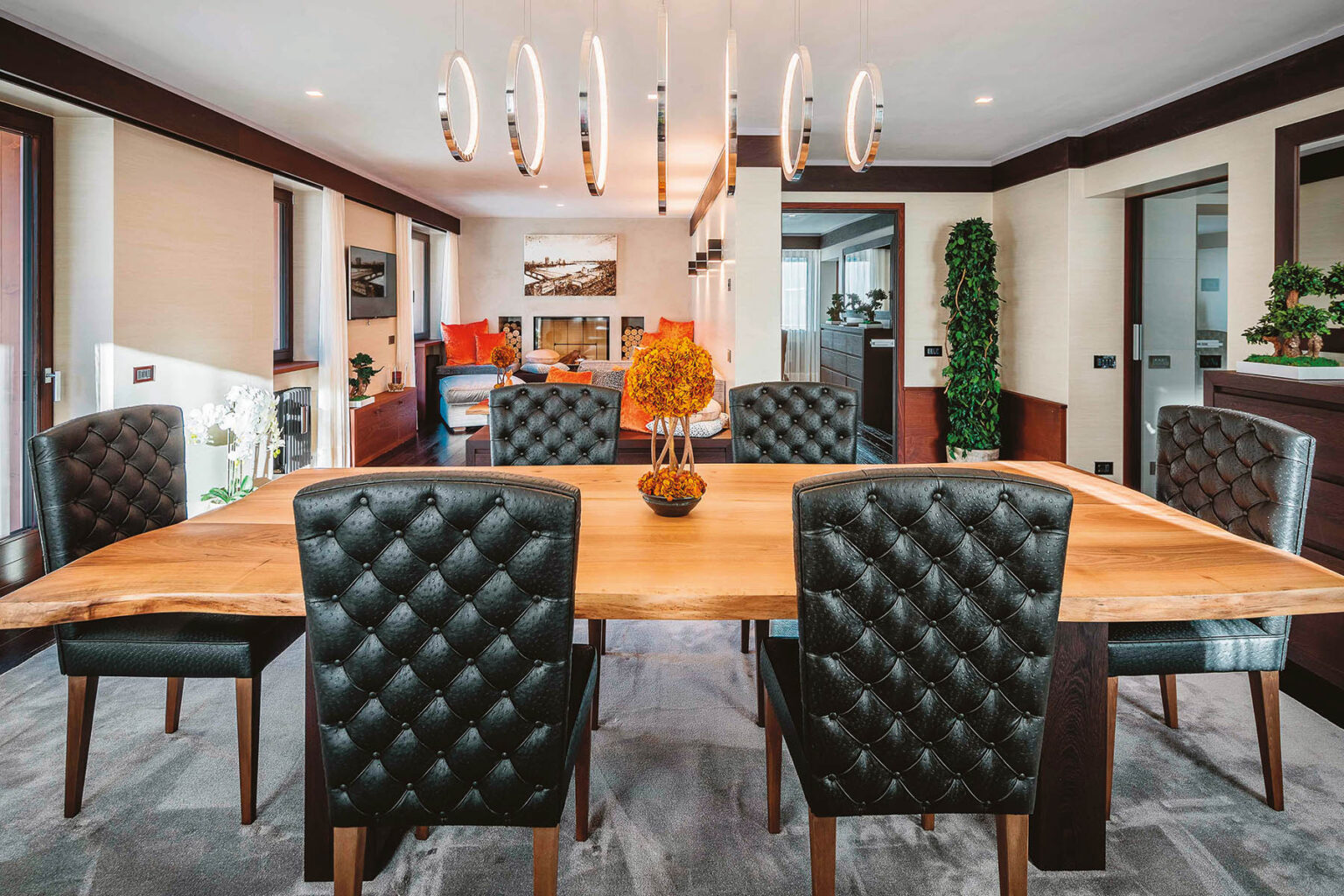 Modern dining room featuring a large wooden table surrounded by tufted black leather chairs, illuminated by a stylish chandelier, with decorative plants in the background. | Only on SopranoVillas