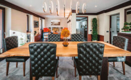Modern dining room featuring a large wooden table surrounded by tufted black leather chairs, illuminated by a stylish chandelier, with decorative plants in the background.