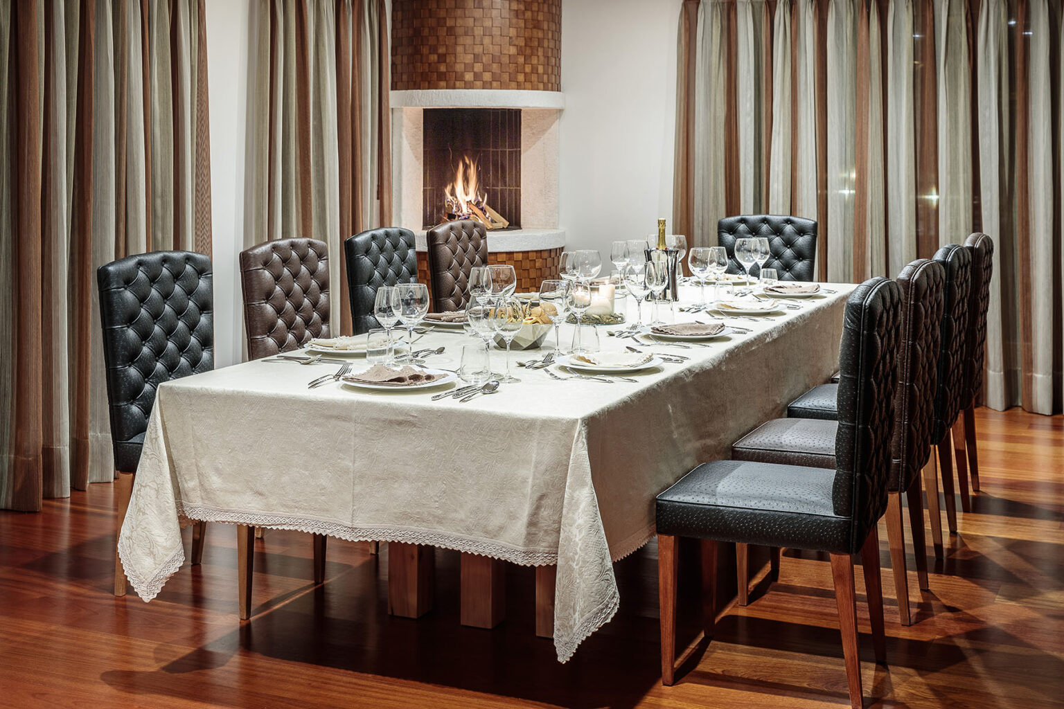 Elegant dining room setup featuring a long table adorned with a white tablecloth, fine china, and glassware. Black and brown tufted chairs surround the table, complemented by soft curtains and a cozy fireplace in the background, creating a warm and inviting atmosphere for gatherings. | Only on SopranoVillas