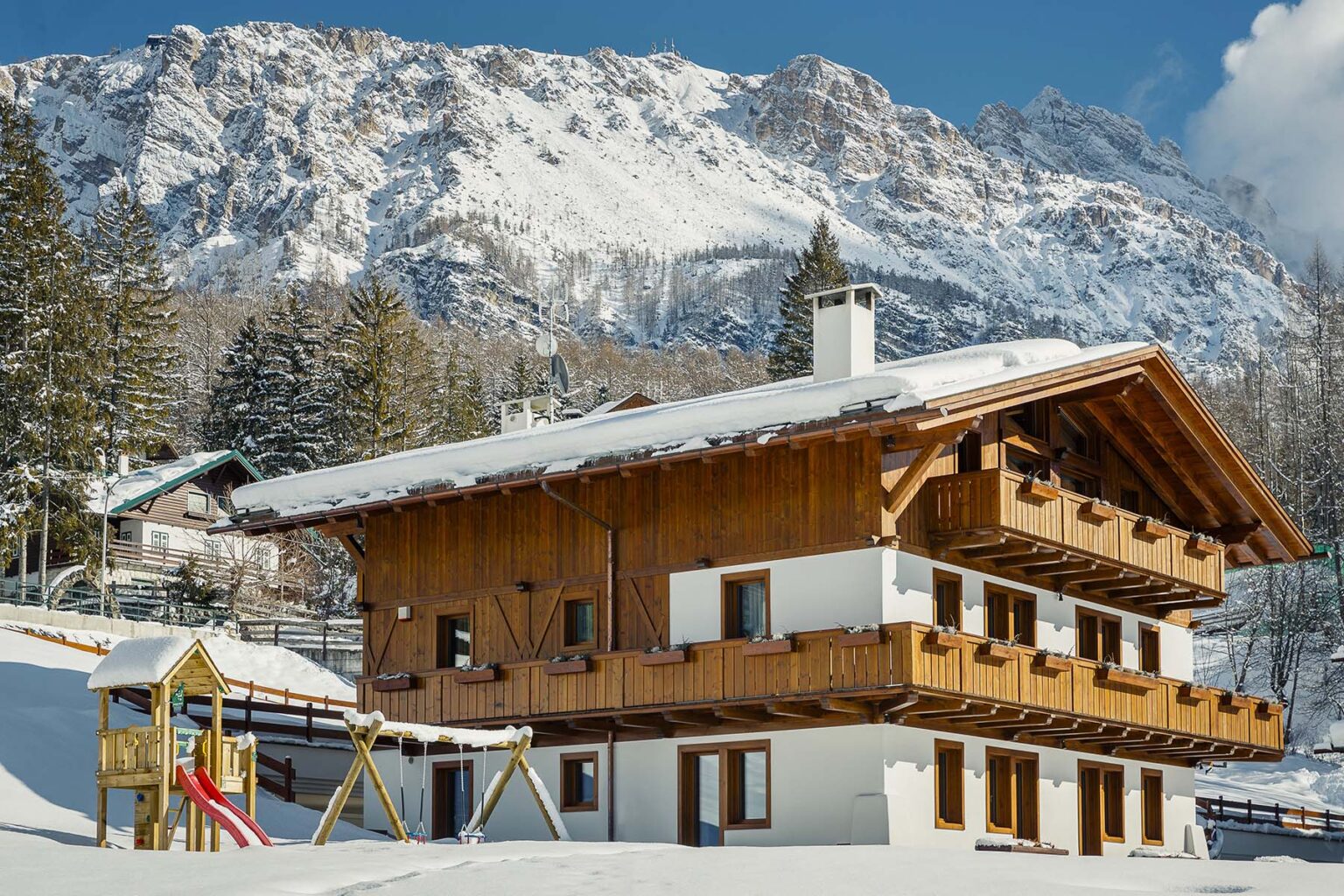 Charming wooden chalet nestled in snow-covered mountains, featuring a playground with a slide and swings in the foreground, surrounded by tall evergreen trees and stunning alpine scenery. | Only on SopranoVillas