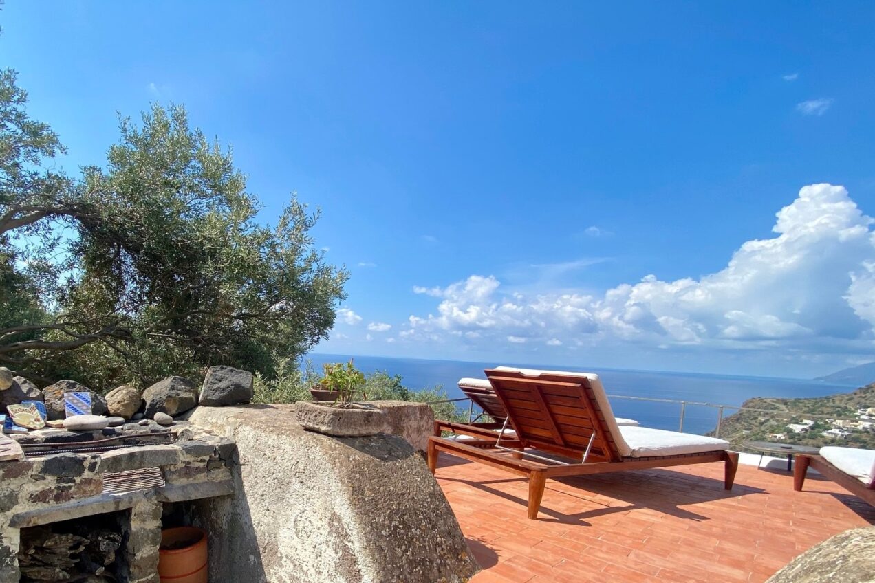 Lounge chairs overlooking a scenic view of the ocean and mountains under a clear blue sky, with an olive tree and stone structure in the foreground. Ideal for relaxation and enjoying nature.