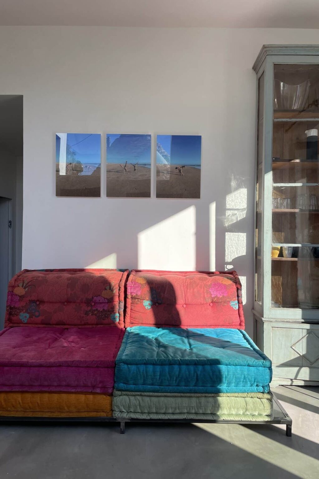 Colorful sofa with pink and blue cushions in a bright room, featuring three beach-themed art pieces on the wall and a wooden cabinet. Natural light casts shadows, enhancing the cozy atmosphere. | Only on SopranoVillas
