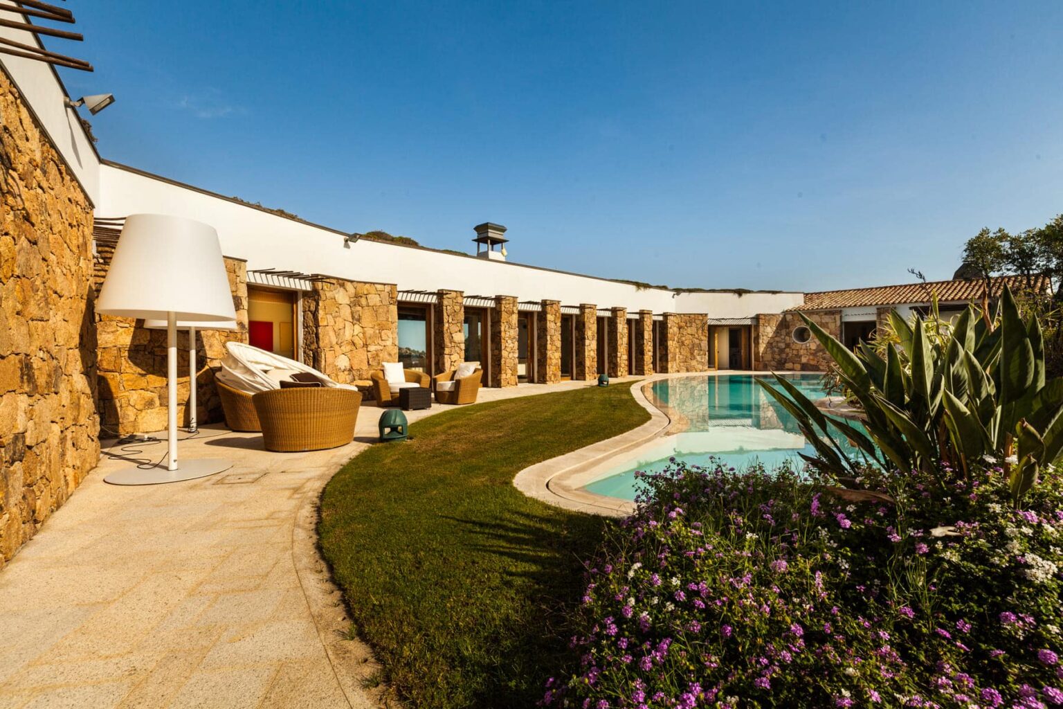 Modern villa with a stone exterior, featuring a landscaped garden, swimming pool, and outdoor seating area. Sunlight illuminates the space, showcasing a stylish lamp and comfortable furniture, perfect for relaxation. | Only on SopranoVillas