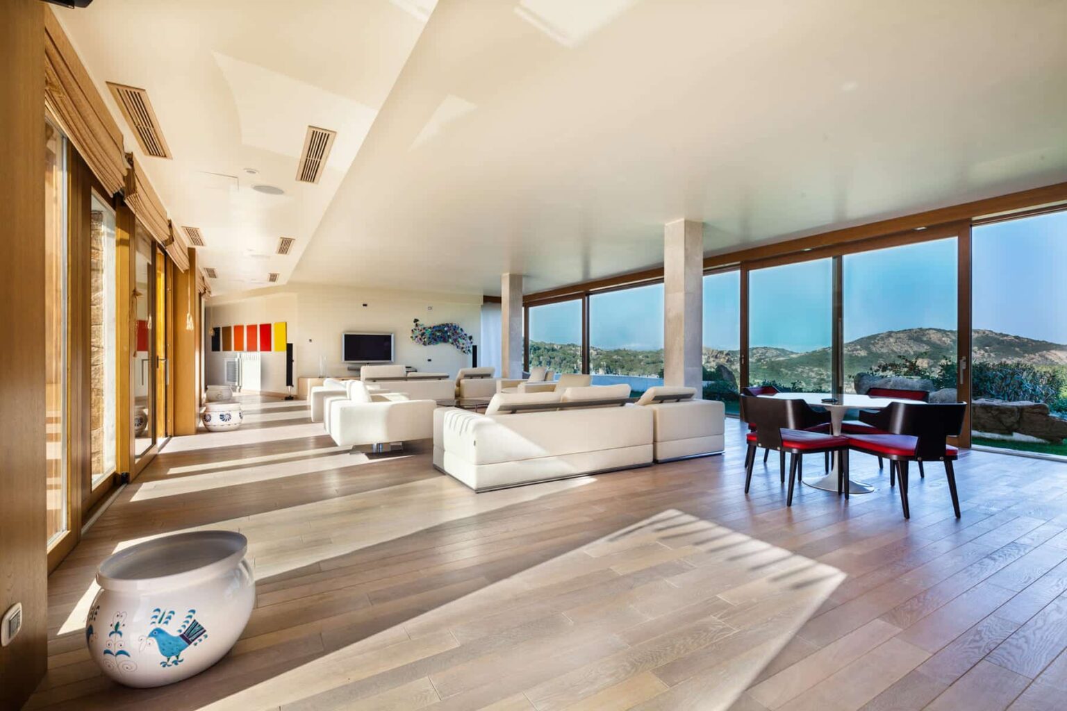 Spacious modern living room with large windows offering scenic views, featuring white sofas, a stylish coffee table, and a dining area with elegant chairs, all set on polished wooden floors. | Only on SopranoVillas