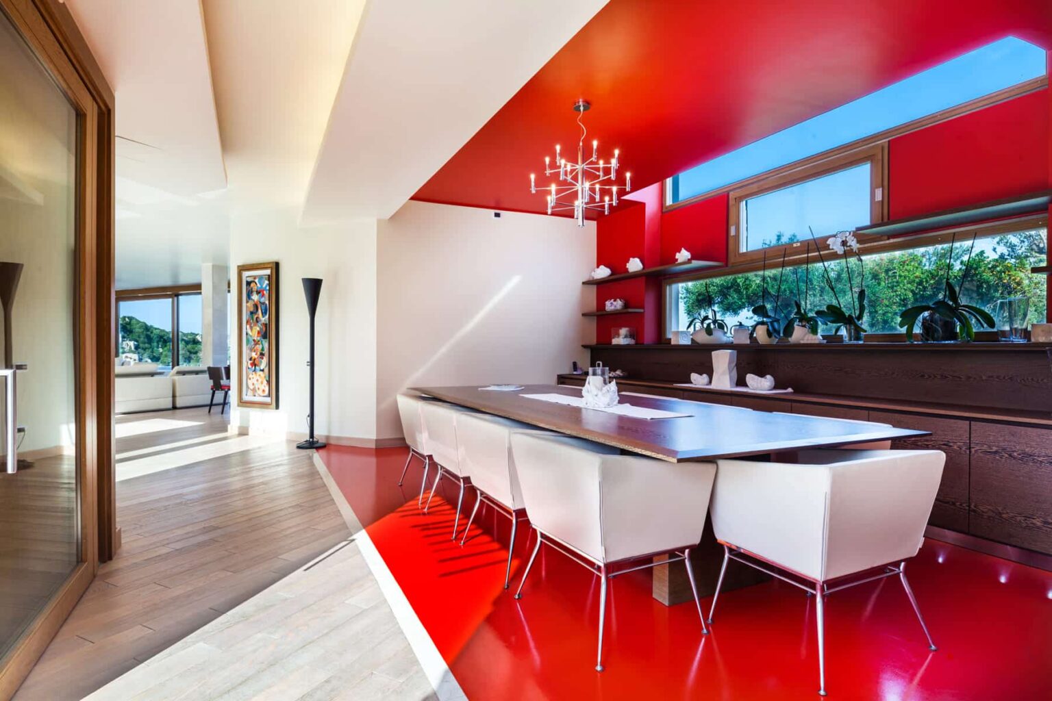 Modern dining room featuring a sleek table surrounded by white chairs, vibrant red walls, and large windows that provide natural light and outdoor views. The design includes contemporary decor elements and a stylish chandelier. | Only on SopranoVillas