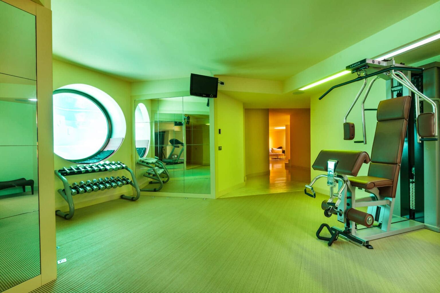 Modern home gym featuring a variety of dumbbells, exercise equipment, and large circular windows, illuminated by green lighting for a vibrant atmosphere. | Only on SopranoVillas