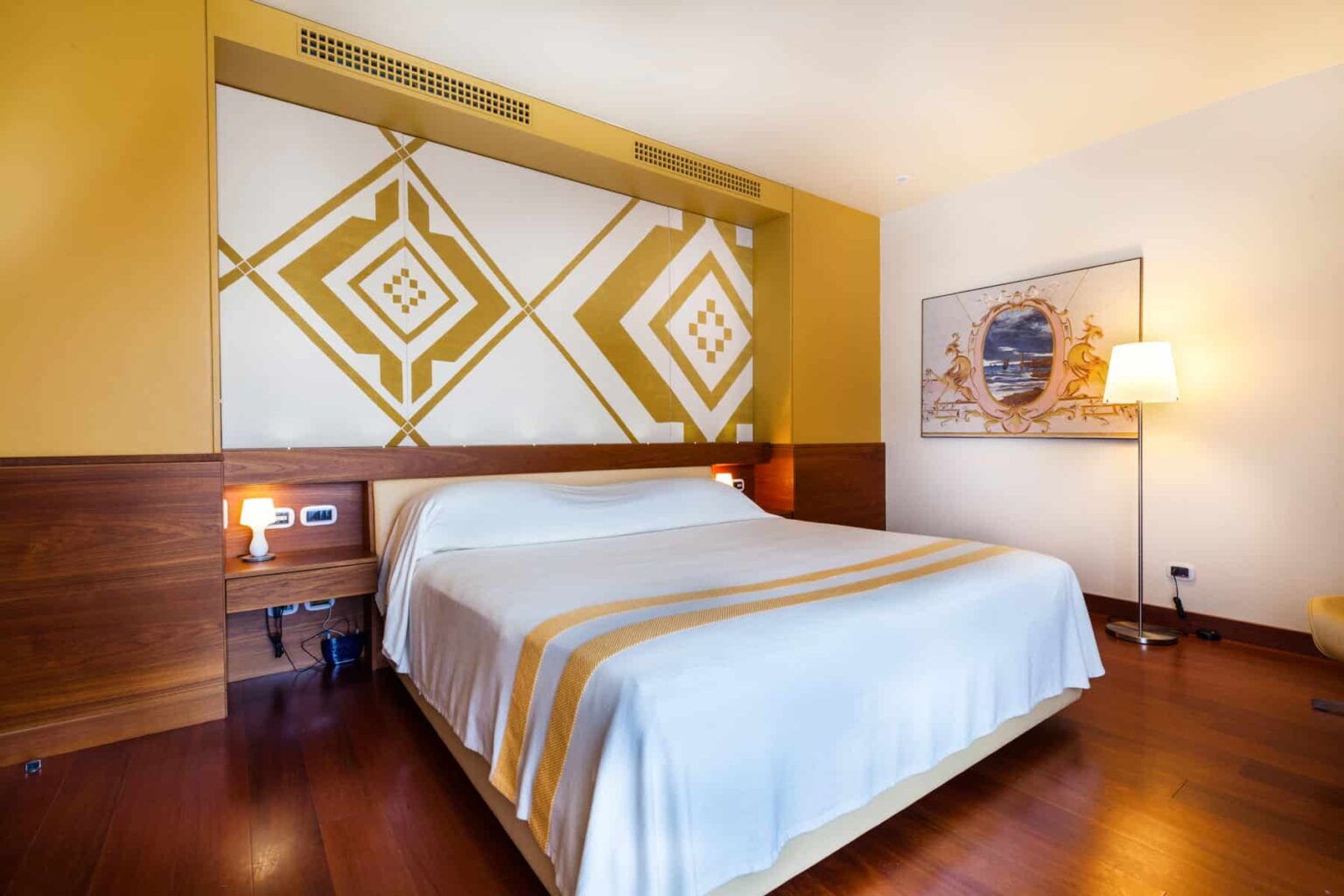 Cozy hotel room featuring a large bed with white linens, wooden accents, and a decorative wall with geometric patterns. The room includes bedside lamps and a framed artwork, creating a warm and inviting atmosphere. Ideal for relaxation and comfort during a stay. | Only on SopranoVillas