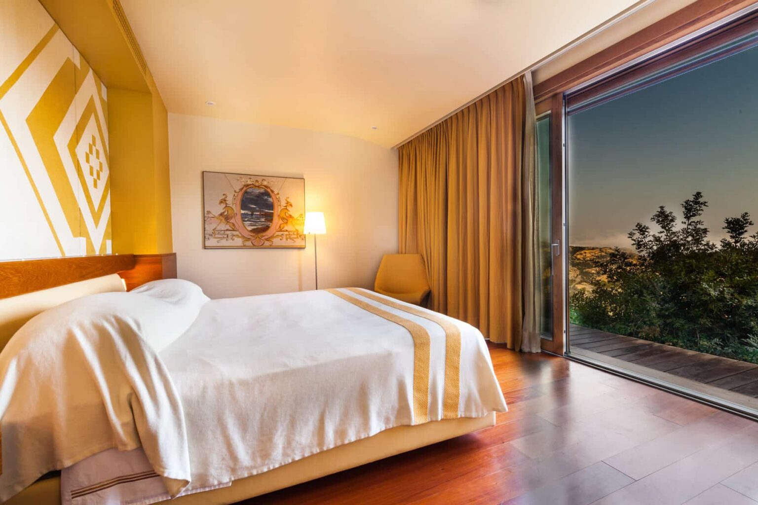 Modern hotel bedroom with a large bed, warm lighting, and a scenic view through a glass door. The room features stylish decor, wooden flooring, and soft curtains, creating a cozy and inviting atmosphere. | Only on SopranoVillas