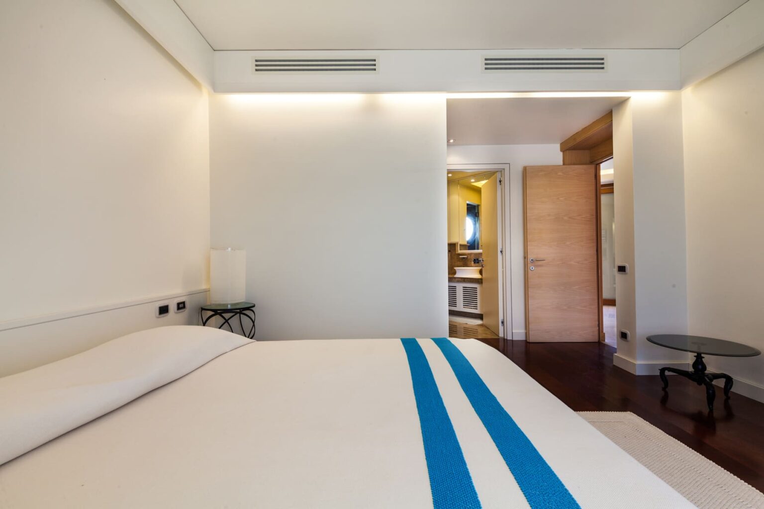 Modern hotel room featuring a neatly made bed with blue striped bedding, minimalist decor, and a doorway leading to a bathroom area, highlighting a spacious and inviting atmosphere. | Only on SopranoVillas