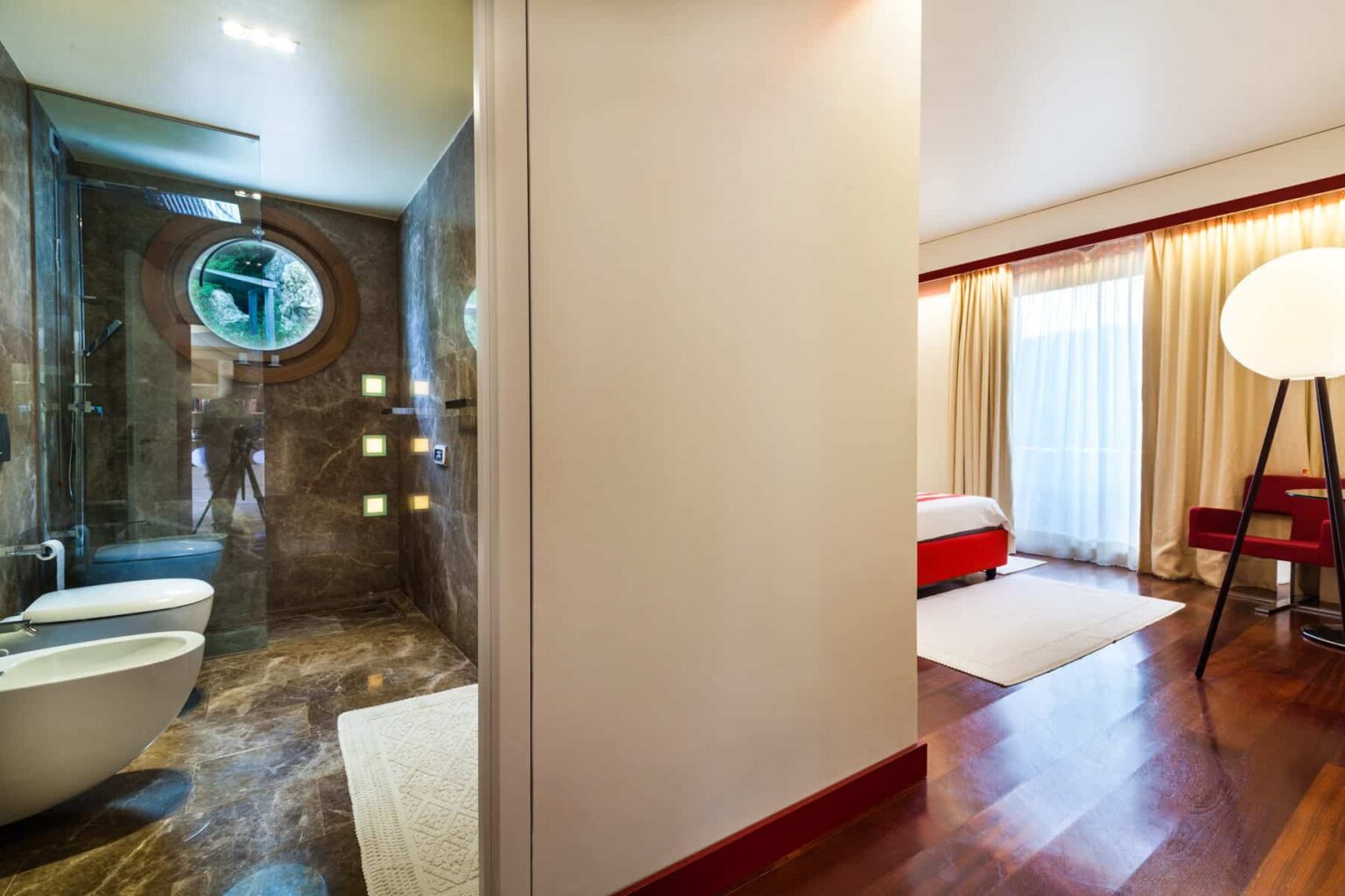 Modern bathroom featuring elegant marble finishes, a glass shower enclosure, and a circular window, with a view into a stylish bedroom decorated in warm tones and natural light. | Only on SopranoVillas